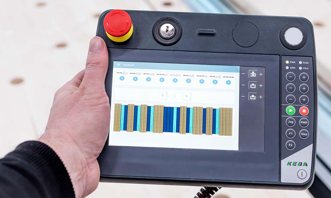 Handheld industrial control panel with a large red emergency stop button and a touchscreen displaying a process interface.