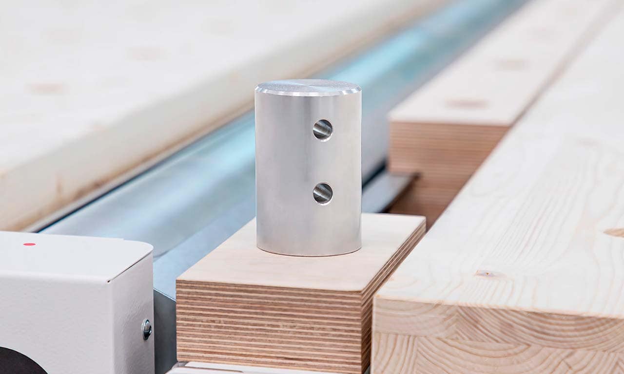 Metal cylindrical weight with two holes sits atop a stack of plywood on an industrial production line