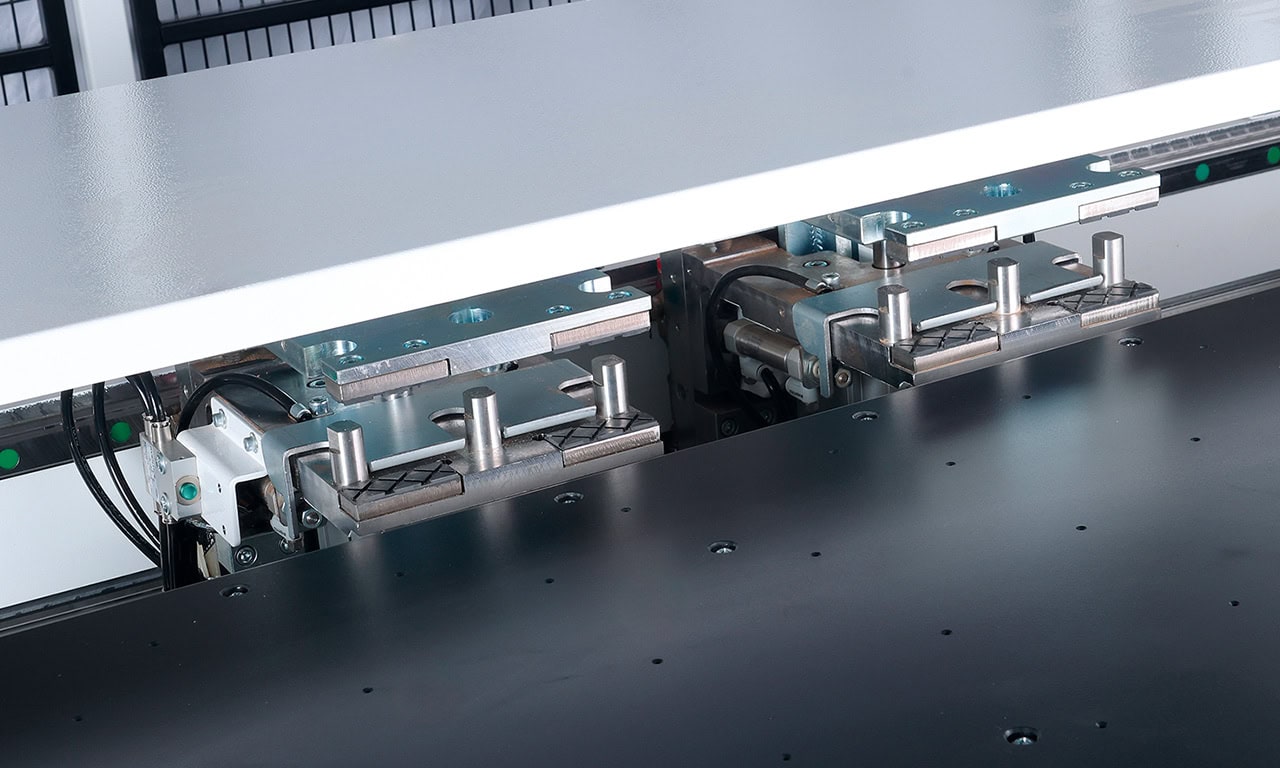 Close-up of a metalworking machine with dual clamp assemblies gripping a white sheet over a dark work surface.