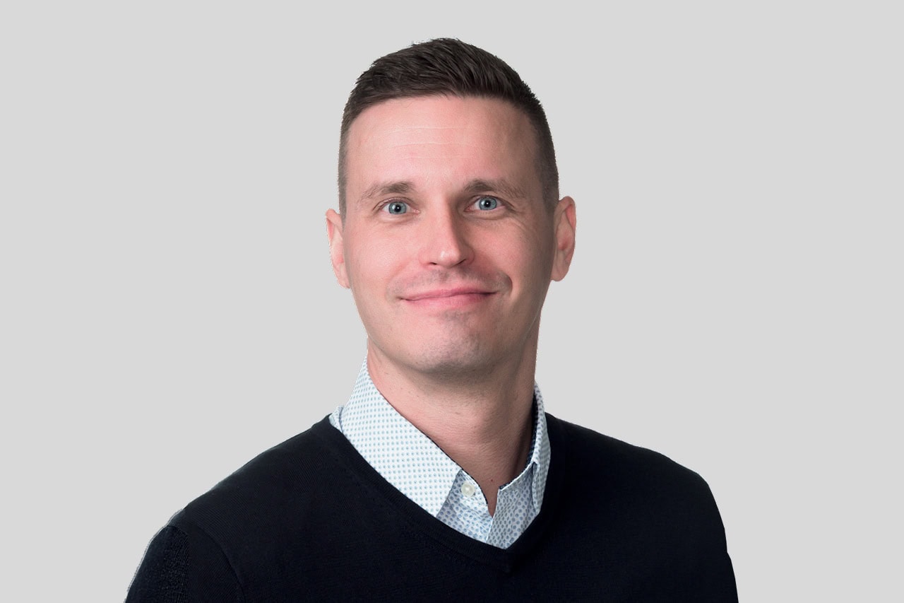 Professional headshot of a smiling man with short dark hair, wearing a light blue button-down under a black sweater, against a light gray background.