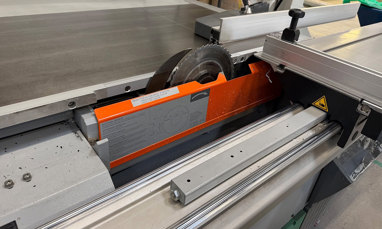 Industrial panel saw with orange safety guard and circular blade, cutting a sheet on a sliding table.