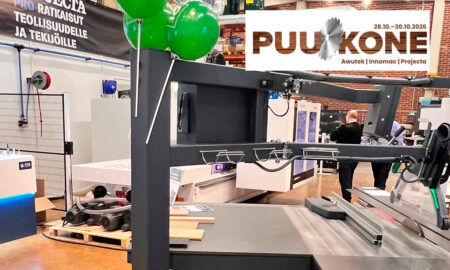 Large metal industrial machine on a display table at a trade show with a PUUKONE banner and green balloons in the background